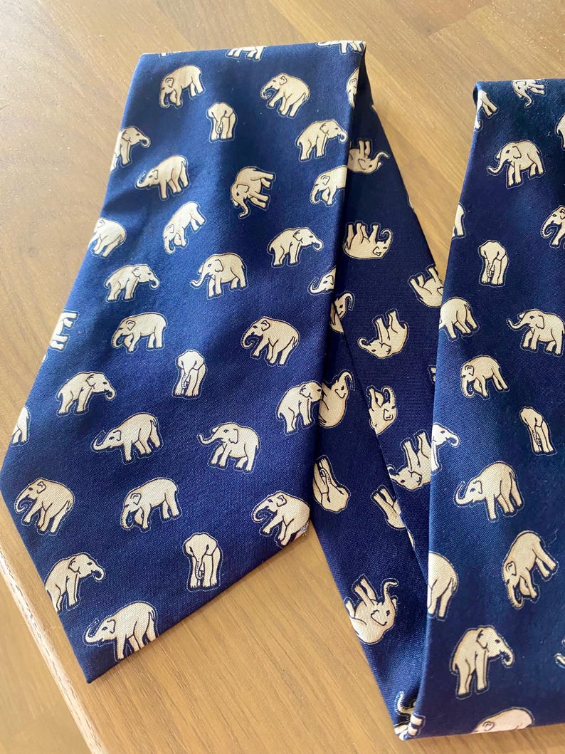 Op de afbeelding: Twee marineblauwe stropdassen met een herhaald patroon van cr&egrave;mekleurige olifanten. De stropdassen zijn gemaakt van een gladde stof en worden gepresenteerd op een lichtgekleurd houten oppervlak. Het olifantenontwerp is gedetailleerd en grillig.