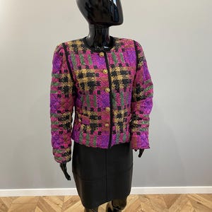 May include: A quilted jacket featuring a vibrant plaid pattern in shades of purple, green, and black, accented with gold-tone buttons down the front and black trim along the collar and placket, paired with a solid black skirt on a mannequin.