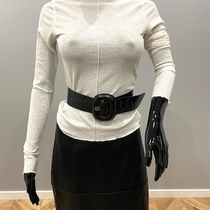 May include: A mannequin displays a cream-colored long-sleeve top with a high neckline, paired with a black leather skirt and a wide black belt with a large buckle. The mannequin's arms are posed, wearing black gloves.