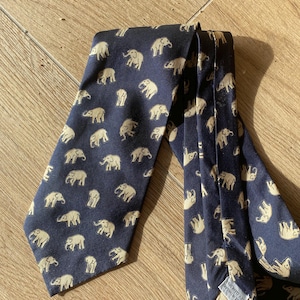 Op de afbeelding: Marineblauwe stropdas met een herhaald patroon van beige olifanten. De stropdas is gemaakt van een gladde stof en is op een houten oppervlak gevouwen. De olifanten zijn in verschillende poses afgebeeld.