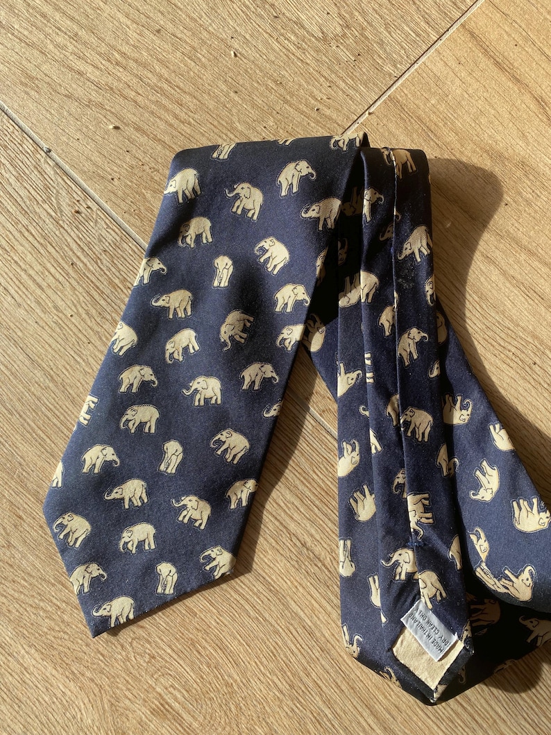 Op de afbeelding: Marineblauwe stropdas met een herhaald patroon van beige olifanten. De stropdas is gemaakt van een gladde stof en is op een houten oppervlak gevouwen. De olifanten zijn in verschillende poses afgebeeld.