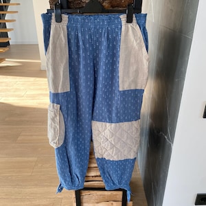 May include: Blue and beige patchwork pants with a textured pattern. The pants have a relaxed fit with beige patches on the front and sides. The bottom of the pants are tied with a string. The pants are hanging on a black hanger.
