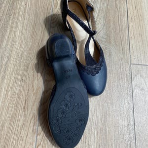 May include: A pair of navy blue T-strap shoes with a low block heel. The shoes feature a textured upper, a scalloped edge detail, and a floral pattern embossed on the dark blue rubber outsole. The brand name Hotter is visible on the insole.