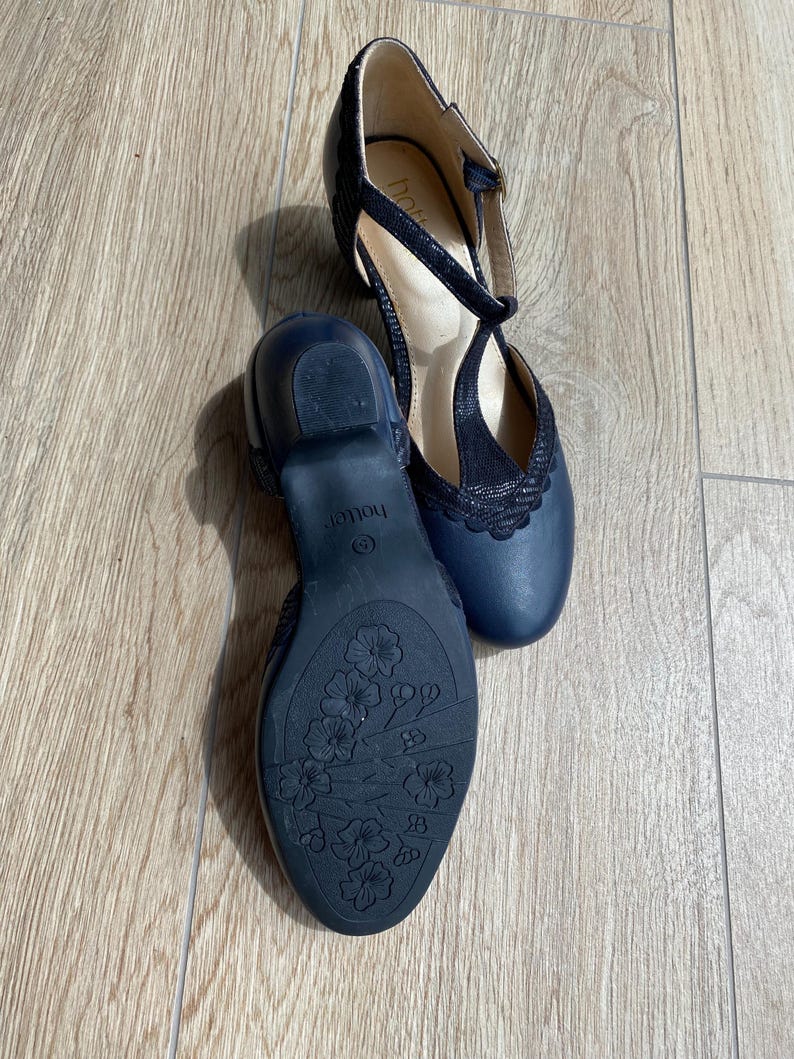 May include: A pair of navy blue T-strap shoes with a low block heel. The shoes feature a textured upper, a scalloped edge detail, and a floral pattern embossed on the dark blue rubber outsole. The brand name Hotter is visible on the insole.