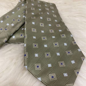 Puede incluir: Corbata verde oliva con un patrón geométrico de líneas diagonales y pequeños cuadrados en azul marino y azul claro. La corbata está hecha de una tela suave, posiblemente seda.
