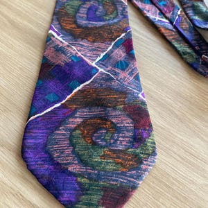 Könnte beinhalten: Eine gemusterte Krawatte mit einem abstrakten Design. Die Krawatte zeigt eine Mischung aus Farben, darunter Lila, Blau, Grün und Orange, mit weißen Akzentlinien. Das Design hat ein wirbelndes Muster und sorgt für visuelles Interesse.