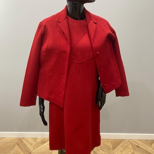 Puede incluir: Un vibrante conjunto vintage rojo de vestido y chaqueta a juego. El vestido llega a la rodilla con un corpiño entallado. La chaqueta presenta un cuello clásico y cierre de botones. El conjunto se exhibe en un maniquí.