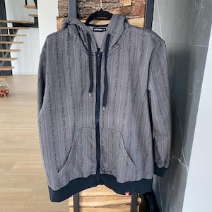 Puede incluir: Una sudadera gris con cremallera y diseño estampado, con capucha, bolsillos delanteros y cremallera negra. La sudadera tiene ribetes negros en los puños y el dobladillo. El texto "ATRIUM 4" es visible en la capucha.