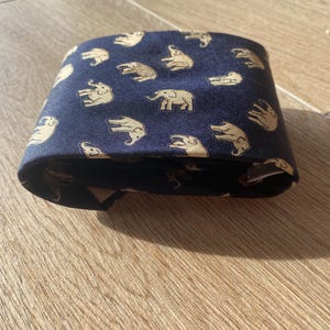 Op de afbeelding: Gevouwen marineblauwe stropdas met een herhalend patroon van beige olifanten. De stropdas is gemaakt van een gladde stof en is netjes opgevouwen, waarbij het olifantenontwerp wordt getoond. De achtergrond is een lichtbruin houten oppervlak.