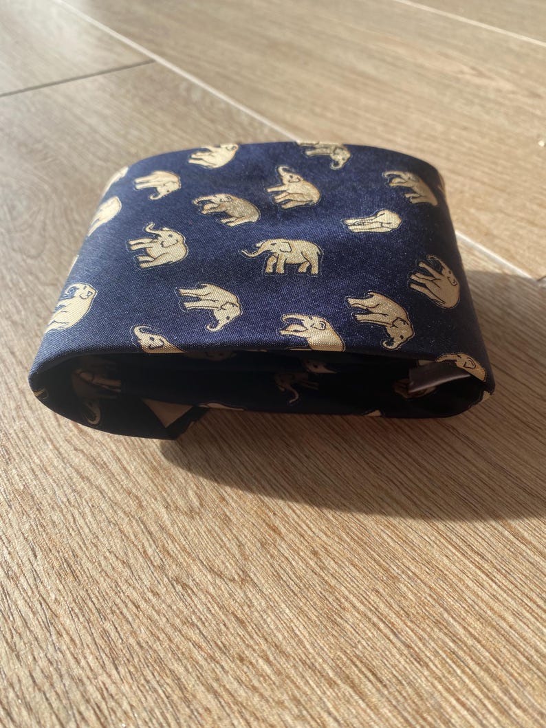 Op de afbeelding: Gevouwen marineblauwe stropdas met een herhalend patroon van beige olifanten. De stropdas is gemaakt van een gladde stof en is netjes opgevouwen, waarbij het olifantenontwerp wordt getoond. De achtergrond is een lichtbruin houten oppervlak.