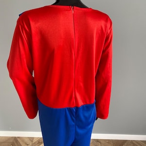 May include: A long-sleeved jumpsuit featuring a vibrant red bodice and bright blue trousers. The garment has a vertical zipper closure running down the center of the back, extending from the neckline to the waist, and is displayed on a mannequin.