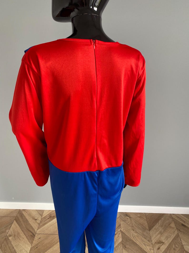 May include: A long-sleeved jumpsuit featuring a vibrant red bodice and bright blue trousers. The garment has a vertical zipper closure running down the center of the back, extending from the neckline to the waist, and is displayed on a mannequin.