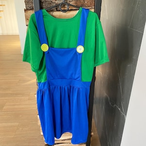 May include: A green t-shirt dress with blue overalls, featuring large yellow buttons. The dress has a gathered skirt and is hanging on a black hanger. The outfit is displayed against a wooden and black metal background.