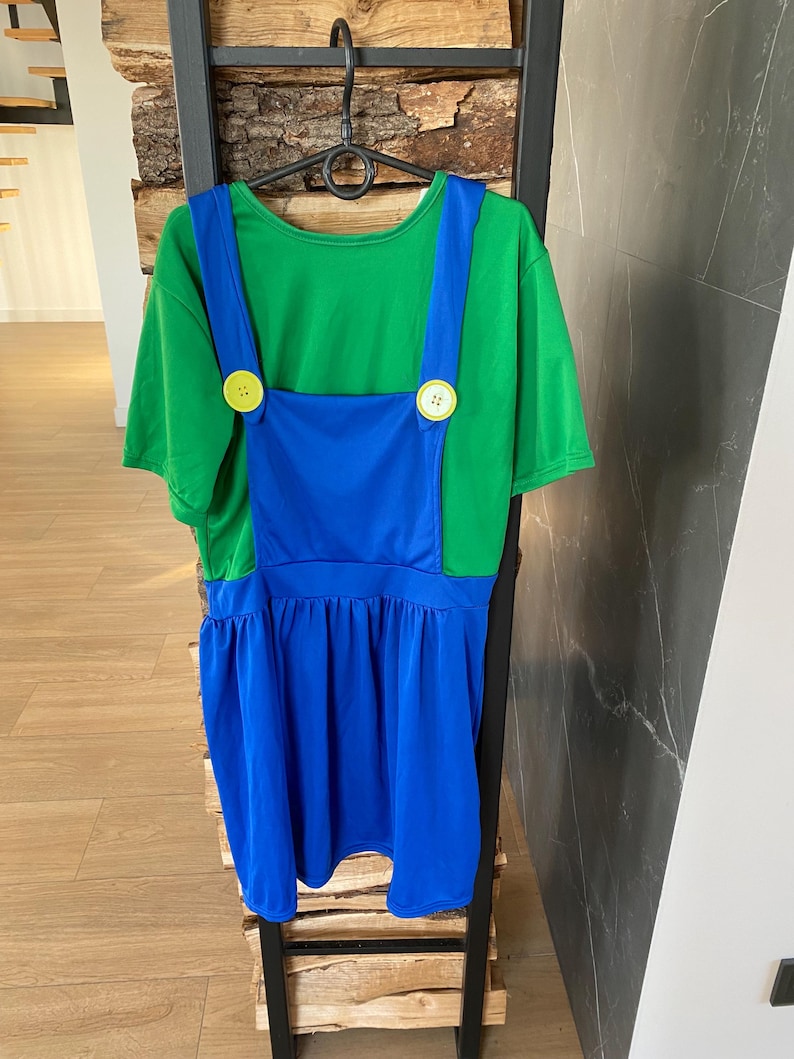 May include: A green t-shirt dress with blue overalls, featuring large yellow buttons. The dress has a gathered skirt and is hanging on a black hanger. The outfit is displayed against a wooden and black metal background.