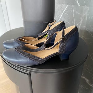 May include: A pair of navy blue T-strap shoes featuring a rounded toe, a low block heel, and a textured black scalloped trim along the collar and strap. The shoes include a thin ankle strap with a small metallic buckle closure and a tan interior lining.