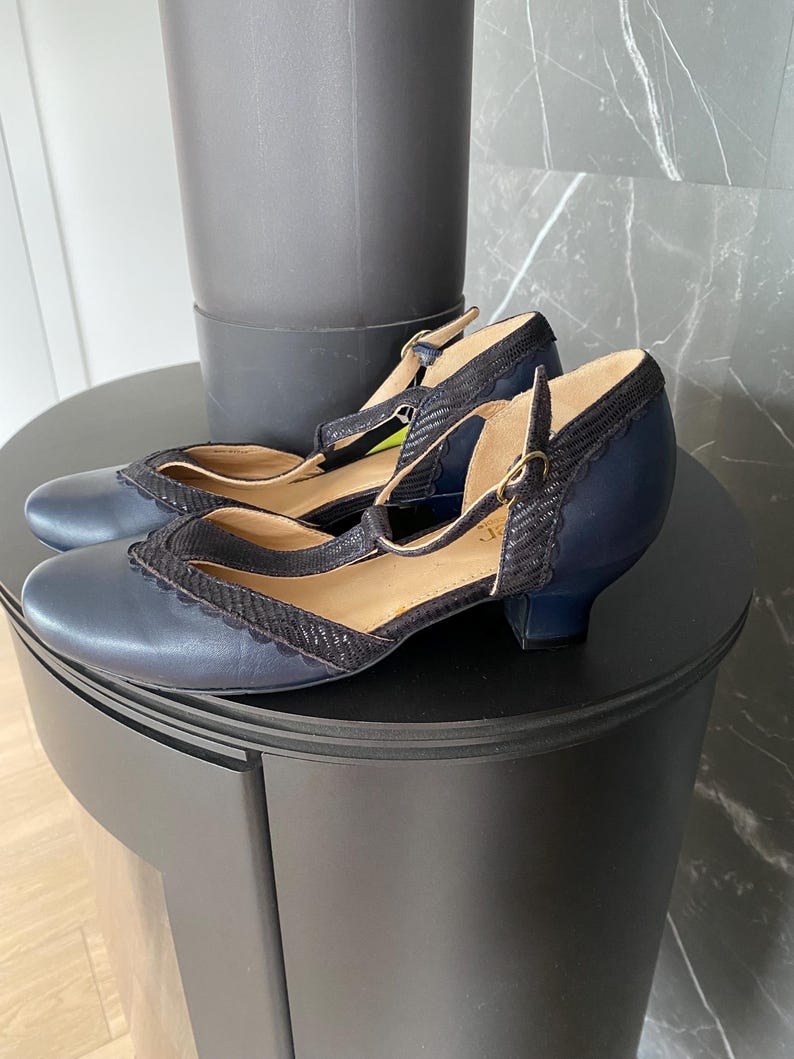 May include: A pair of navy blue T-strap shoes featuring a rounded toe, a low block heel, and a textured black scalloped trim along the collar and strap. The shoes include a thin ankle strap with a small metallic buckle closure and a tan interior lining.