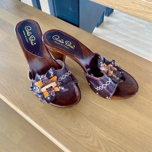 May include: A pair of brown wooden heeled sandals with floral detailing. The sandals feature a dark brown leather upper with decorative stitching and a wooden heel. The brand name "Carlo Pera" is visible on the insole.