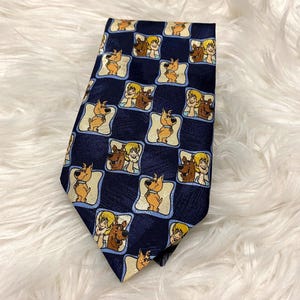 May include: A navy blue necktie featuring a repeating pattern of cartoon characters. The pattern includes images of a tan dog and two people with blonde hair. The tie has a checkered design with the cartoon characters in each square.