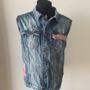 May include: A distressed denim vest with patches of the American flag. The vest has a button-up front, pockets, and a collared neckline. The denim is faded and has some rips and tears for a vintage look.
