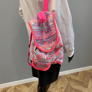 May include: A backpack featuring a vibrant paisley pattern in shades of pink, blue, and white. The bag is accented with bright neon pink pom-pom trim along the edges of the flap and front pocket, and includes matching neon pink shoulder straps.