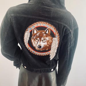 Puede incluir: Chaqueta de pana negra con un gran parche bordado con la cabeza de un lobo, plumas y un diseño circular. La chaqueta tiene una parte delantera abotonada y un cuello clásico. El parche es marrón, beige y blanco.