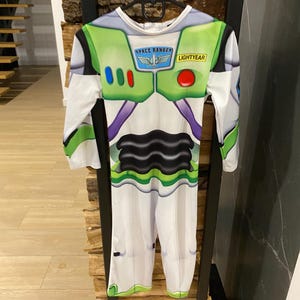 May include: A Buzz Lightyear costume with long sleeves and pants. The white jumpsuit features green and purple accents, with the words "SPACE RANGER" and "LIGHTYEAR" printed on the chest. The costume is hanging on a black hanger.