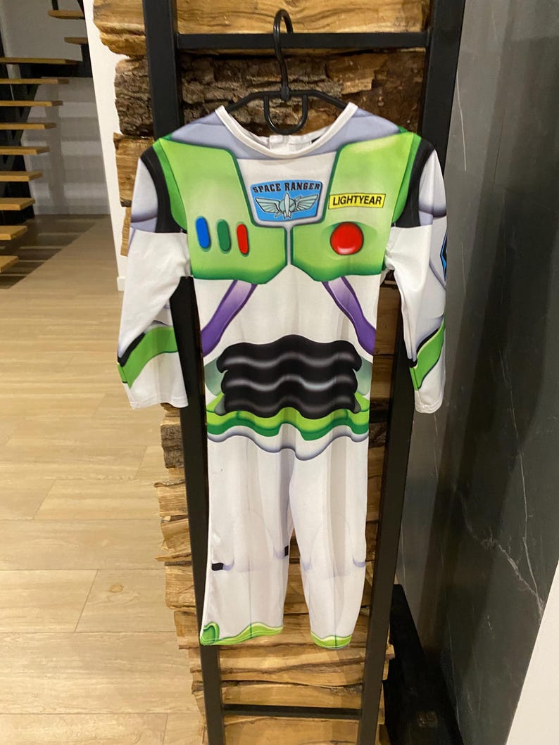 May include: A Buzz Lightyear costume with long sleeves and pants. The white jumpsuit features green and purple accents, with the words "SPACE RANGER" and "LIGHTYEAR" printed on the chest. The costume is hanging on a black hanger.