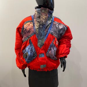 Puede incluir: Una chaqueta de esquí roja vintage con paneles estampados coloridos en los hombros y el pecho. La chaqueta tiene un cuello alto y detalles de botones. La chaqueta se exhibe en un maniquí negro.