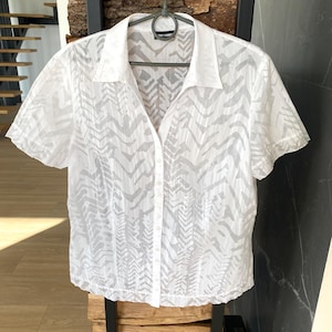 Pode incluir: Camisa branca de manga curta com um padrão texturizado sutil. A camisa tem uma gola clássica e está pendurada em um cabide de metal. O fundo inclui elementos de madeira e cinza escuro.