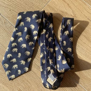 Op de afbeelding: Marineblauwe stropdas met een herhaald patroon van beige olifanten. De stropdas is gemaakt van een gladde stof en wordt gepresenteerd op een houten oppervlak. De olifanten zijn gelijkmatig verdeeld over de stropdas.