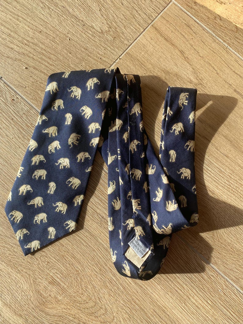 Op de afbeelding: Marineblauwe stropdas met een herhaald patroon van beige olifanten. De stropdas is gemaakt van een gladde stof en wordt gepresenteerd op een houten oppervlak. De olifanten zijn gelijkmatig verdeeld over de stropdas.