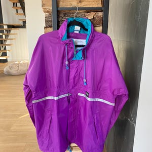 Könnte beinhalten: Eine leuchtend violette Vintage-Jacke mit türkisfarbenem Kragen und Akzenten. Die Jacke hat eine Kapuze mit Kordelzug, reflektierende Streifen und einen Reißverschluss. Die Jacke hängt an einem schwarzen Gestell.