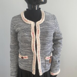 Può includere: Giacca in maglia grigia con finiture color crema e dettagli rossi. La giacca ha due tasche anteriori con bottoni marroni e finiture color crema. La giacca è esposta su un manichino nero.
