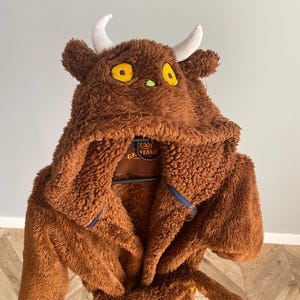 Puede incluir: Bata de felpa marrón con un diseño del personaje Gruffalo. La bata tiene una capucha con cuernos blancos, ojos amarillos y un cinturón. La bata está hecha de un material suave y difuso.