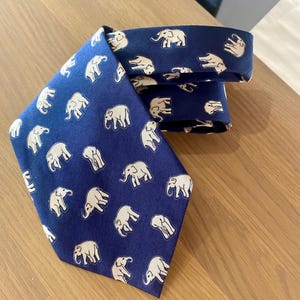 Op de afbeelding: Marineblauwe stropdas met een herhaald patroon van beige olifanten. De stropdas is gemaakt van een gladde stof en ligt op een houten oppervlak. De olifanten zijn afgebeeld in een eenvoudig, gestileerd ontwerp.