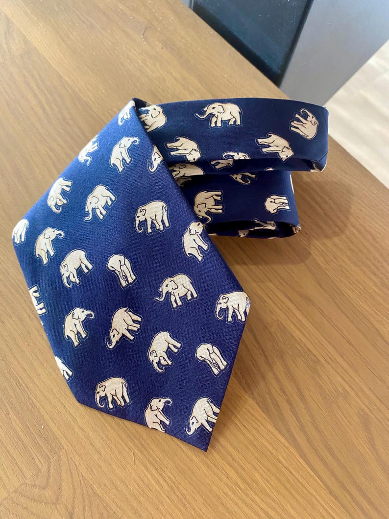 Op de afbeelding: Marineblauwe stropdas met een herhaald patroon van beige olifanten. De stropdas is gemaakt van een gladde stof en ligt op een houten oppervlak. De olifanten zijn afgebeeld in een eenvoudig, gestileerd ontwerp.
