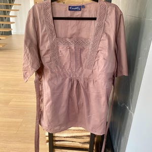 May include: A dusky pink blouse with a square neckline and short sleeves. The neckline and upper chest have a delicate lace trim. The blouse has a gathered waist and long ties. The label reads "Casa & Co."