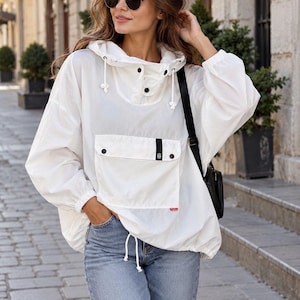 Könnte beinhalten: Eine weiße Windjacke mit Kapuze, die eine große Fronttasche mit Klappe und schwarzen Knopfverschlüssen, eine Kapuze mit Kordelzug, elastische Ärmelbündchen und einen Kordelzug am Saum aufweist. Dazu werden blaue Jeans und eine schwarze Umhängetasche getragen.