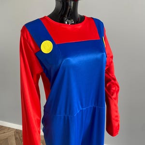May include: A long-sleeved jumpsuit featuring a red shirt base with an attached blue overall overlay. The blue section includes two prominent yellow circular buttons positioned on the chest straps, creating a distinct color-blocked appearance.