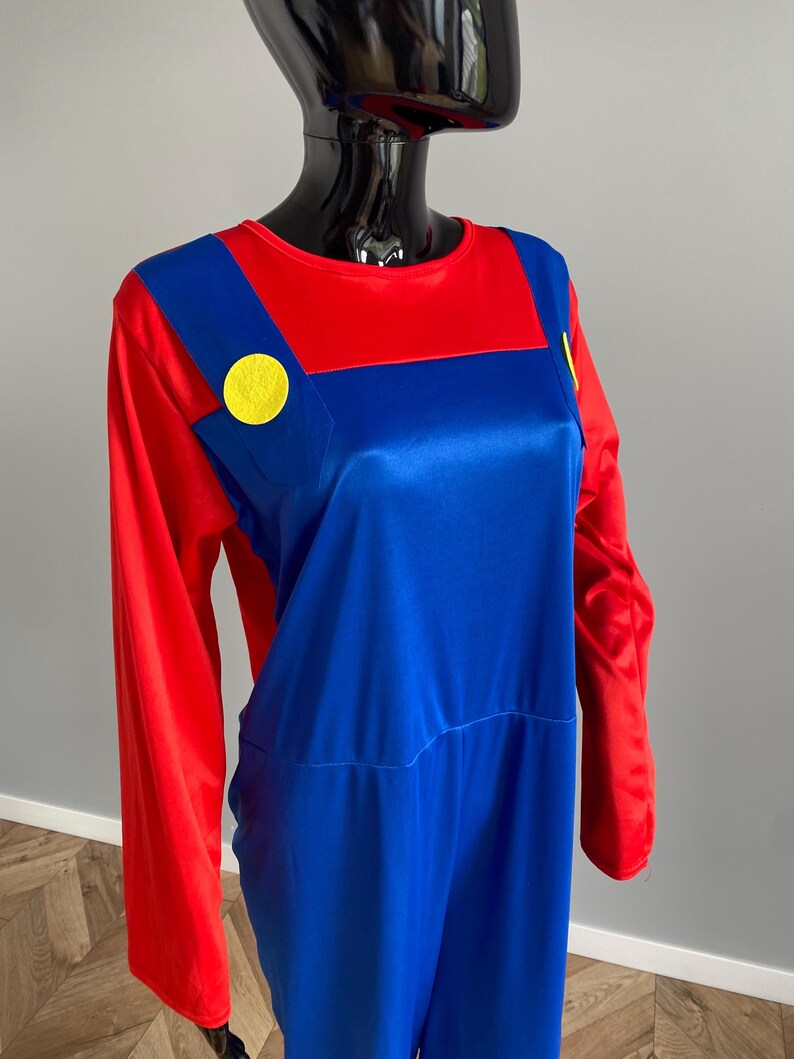 May include: A long-sleeved jumpsuit featuring a red shirt base with an attached blue overall overlay. The blue section includes two prominent yellow circular buttons positioned on the chest straps, creating a distinct color-blocked appearance.