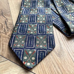Può includere: Una cravatta blu navy con un motivo ripetuto di mazze da golf e palline da golf in un motivo a griglia. Le mazze da golf sono rosse e bianche e le palline da golf sono bianche. Lo sfondo è verde tenue.