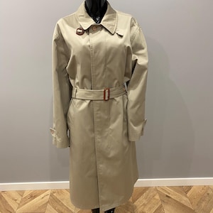 May include: A tan trench coat with a belt, collar, and buttoned cuffs. The coat is displayed on a mannequin. The belt has a brown buckle. The coat is a neutral color and appears to be made of a durable fabric.