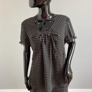 May include: Black blouse with white polka dots, displayed on a mannequin. The blouse features a V-neckline, short sleeves with ruffled edges, and a gathered detail at the bust. The fabric appears lightweight and flowing, suitable for casual wear.
