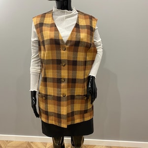 May include: A vintage-style, button-down vest in a plaid pattern of brown, tan, and gold. The vest has two front pockets and is displayed on a mannequin. The mannequin is wearing a white turtleneck and a black skirt.