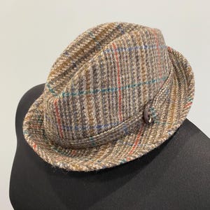 Könnte beinhalten: Ein brauner Tweedhut mit einem rot, blau und grün karierten Muster. Der Hut hat eine kleine Krempe und einen Knopf an der Seite. Ein stilvolles Accessoire.