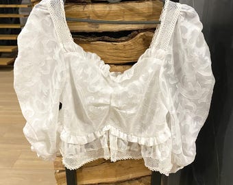May include: A white, semi-sheer blouse with a square neckline and short, puffy sleeves. The blouse features a delicate floral pattern and is trimmed with lace along the neckline, sleeves, and hem. It hangs on a gray hanger.