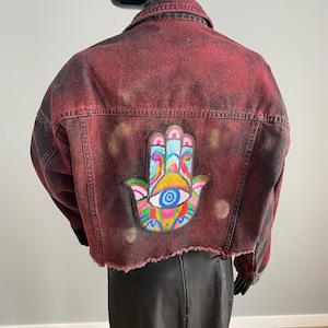 以下が含まれることがあります： 赤色のデニムジャケット、ユーズド加工。 ジャケットの背面には、カラフルなビーズのハムサハンドパッチと目のデザインが施されています。 ジャケットは、切りっぱなしの裾とクラシックな襟が特徴です。