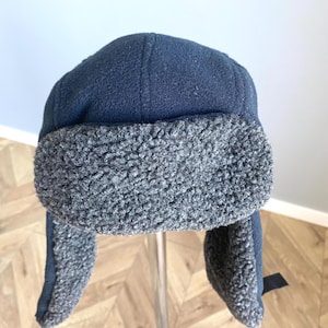May include: A navy blue winter hat with ear flaps and a gray, fuzzy lining. The hat is made of a soft, textured fabric and is designed to keep the wearer warm in cold weather. The ear flaps can be tied under the chin.