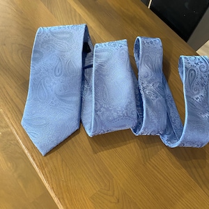 Puede incluir: Una corbata azul claro con estampado de cachemira, exhibida sobre una superficie de madera. La corbata está hecha de un material sedoso y está cuidadosamente doblada y dispuesta, mostrando su diseño y color.
