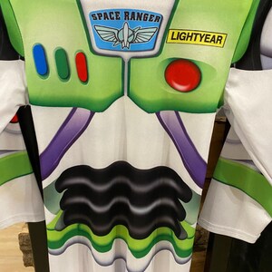 May include: A Buzz Lightyear costume featuring a white and green design with the words "SPACE RANGER" and "LIGHTYEAR". The costume has a green collar and accents, with a red button and three colored buttons on the chest.